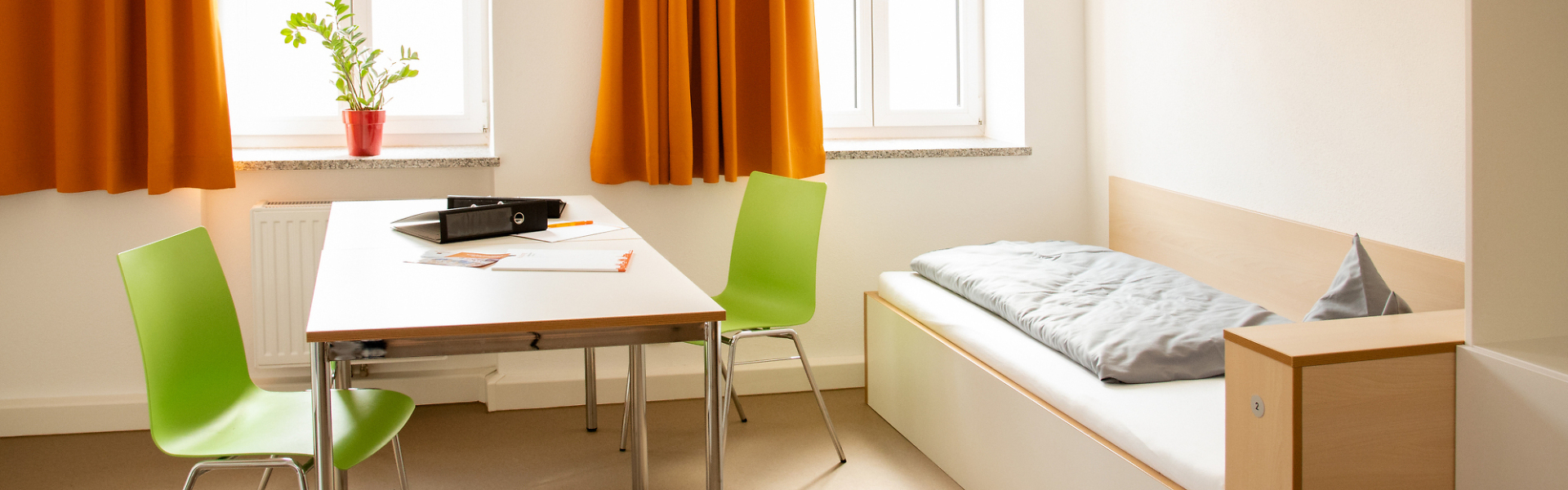 Einfach eingerichtetes Zimmer mit Bett, Tisch, zwei Stühlen und großen Fenstern mit orangefarbenen Vorhängen.