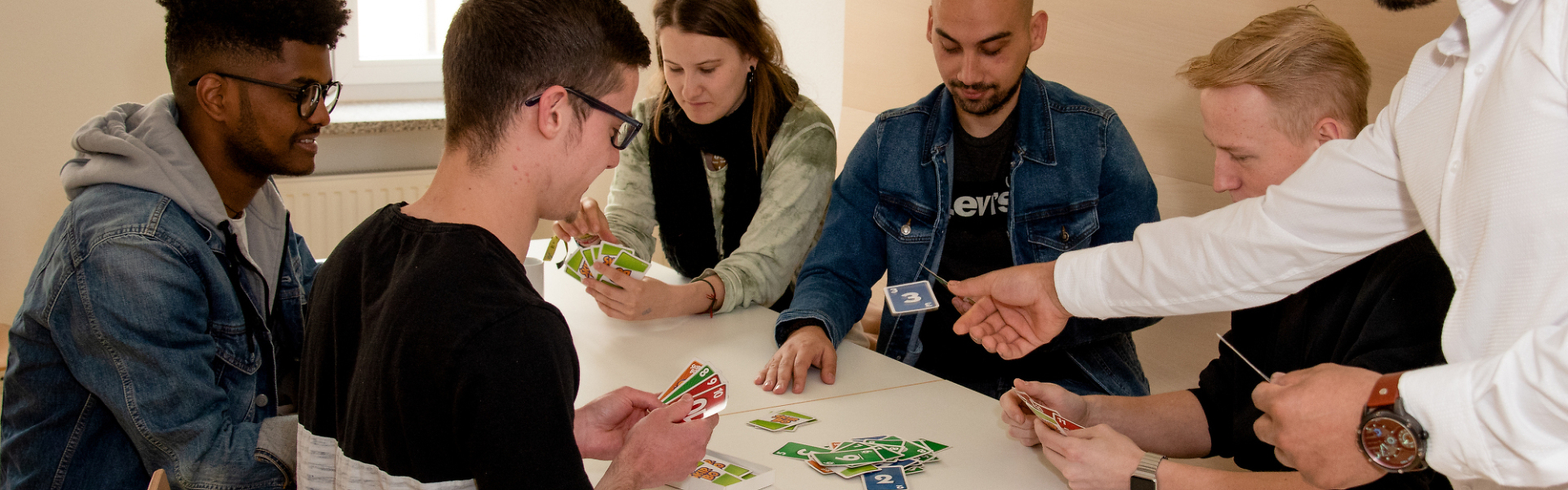 Gruppe junger Erwachsener sitzt gemeinsam an einem Tisch und spielt ein Kartenspiel in einem hellen Gemeinschaftsraum.