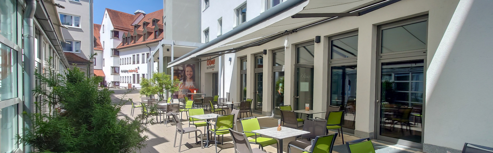 Außenterrasse mit Tischen und grünen Stühlen vor einem Gebäude.