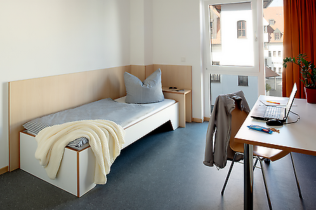 Modernes Einzelzimmer mit Bett, Schreibtisch, Laptop und Fenster mit Blick nach draußen.
