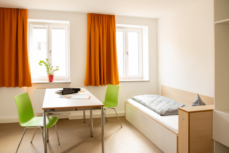 Einfach eingerichtetes Zimmer mit Bett, Tisch, zwei Stühlen und großen Fenstern mit orangefarbenen Vorhängen.