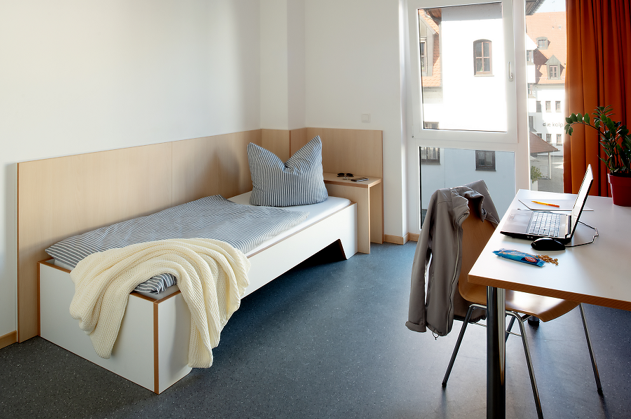 Modernes Einzelzimmer mit Bett, Schreibtisch, Laptop und Fenster mit Blick nach draußen.