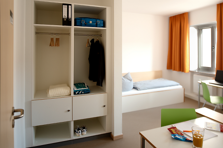 Zimmer mit offenem Kleiderschrank, Bett und Schreibtisch.