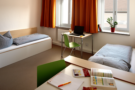 Helles Zweibettzimmer mit Schreibtisch, Laptop und orangefarbenen Vorhängen.