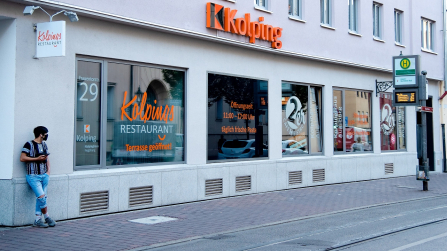 Straßenansicht eines Gebäudes mit Kolping Logo und großer Fensterfront mit Beschriftung und Bushaltestelle davor.