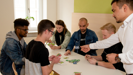 Gruppe junger Erwachsener sitzt gemeinsam an einem Tisch und spielt ein Kartenspiel in einem hellen Gemeinschaftsraum.