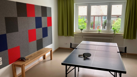 Tischtennisraum mit Platte, Schlägern und großer Fensterfront in hellem Gemeinschaftsraum.